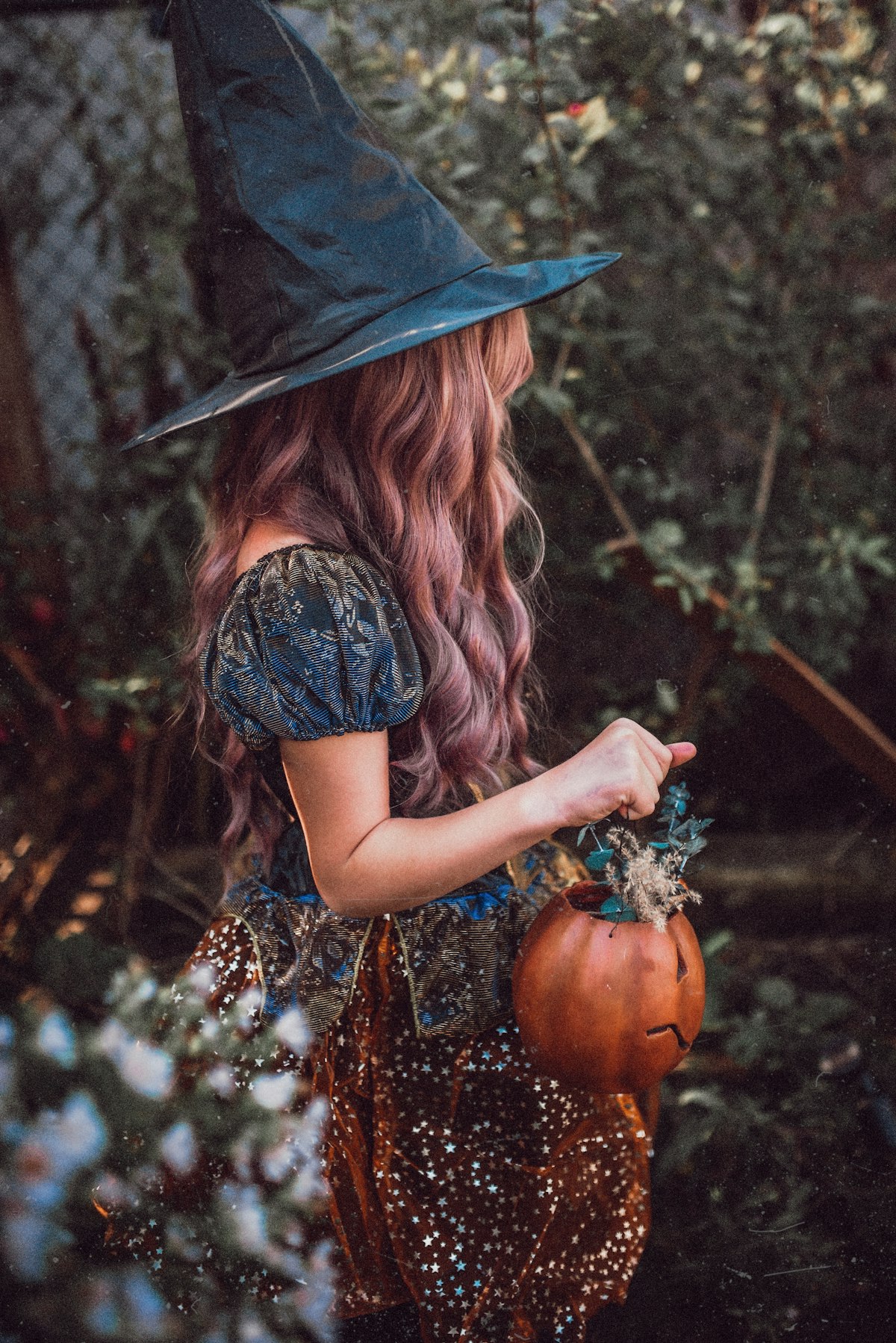 A person in a witch hat and everyday clothes holding a pumpkin bucket