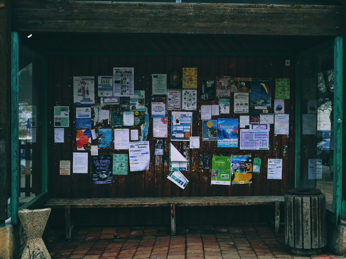 A community noticeboard covered in event flyers beside a wooden bench